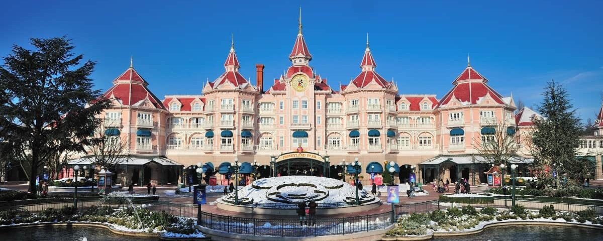 La vue de face de l'hôtel Disneyland Paris avec sa façade rose et blanche élaborée, avec des tourelles et des détails architecturaux complexes. Au premier plan, une fontaine avec des figures de cygnes sous un ciel bleu clair.