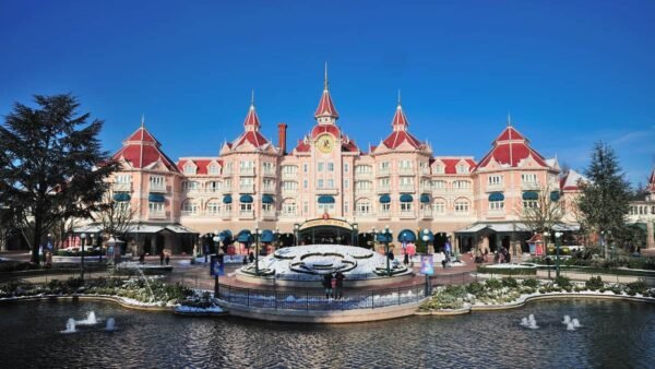 La vue de face de l'hôtel Disneyland Paris avec sa façade rose et blanche élaborée, avec des tourelles et des détails architecturaux complexes. Au premier plan, une fontaine avec des figures de cygnes sous un ciel bleu clair.