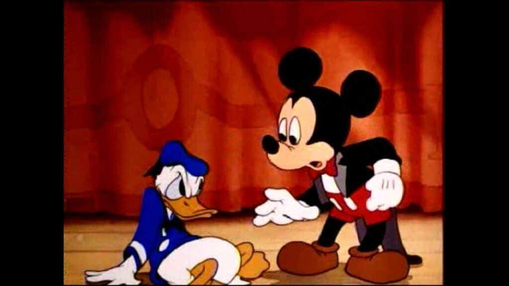Mickey Mouse, dans son short rouge classique et ses gants blancs, semble consoler Donald Duck, qui a l'air bouleversé et est assis par terre devant un fond marron uni.