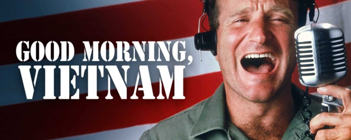 Un homme joyeux portant des écouteurs parle avec animation dans un microphone vintage, avec en toile de fond le drapeau américain et le texte en gras « Good Morning, Vietnam.
