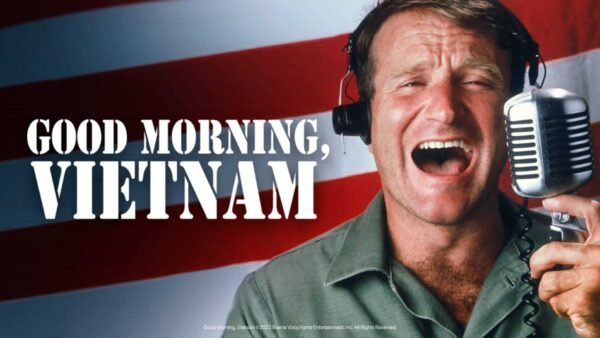 Un homme joyeux portant des écouteurs parle avec animation dans un microphone vintage, avec en toile de fond le drapeau américain et le texte en gras « Good Morning, Vietnam.