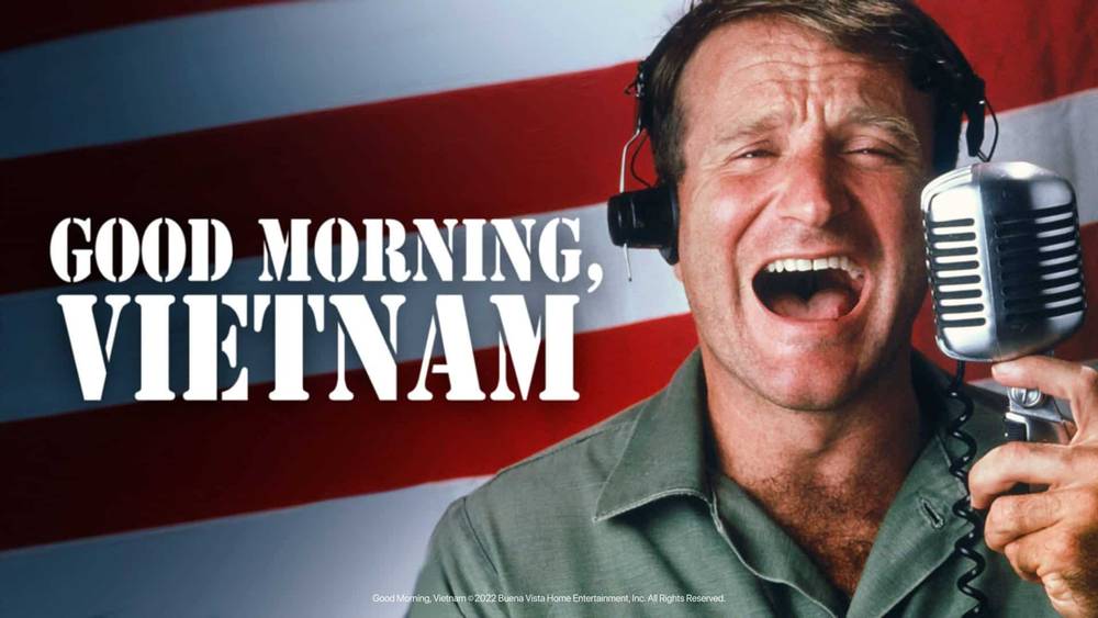 Un homme joyeux portant des écouteurs parle avec animation dans un microphone vintage, avec en toile de fond le drapeau américain et le texte en gras « Good Morning, Vietnam.
