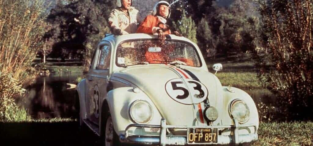 Une scène classique du film "La Coccinelle à Monte Carlo" : deux hommes en tenue de course sont assis au sommet d'une Volkswagen Beetle blanche, numéro 53, connue sous le nom de Herbie, dans