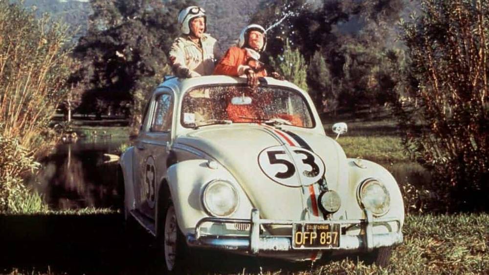 Une scène classique du film "La Coccinelle à Monte Carlo" : deux hommes en tenue de course sont assis au sommet d'une Volkswagen Beetle blanche, numéro 53, connue sous le nom de Herbie, dans