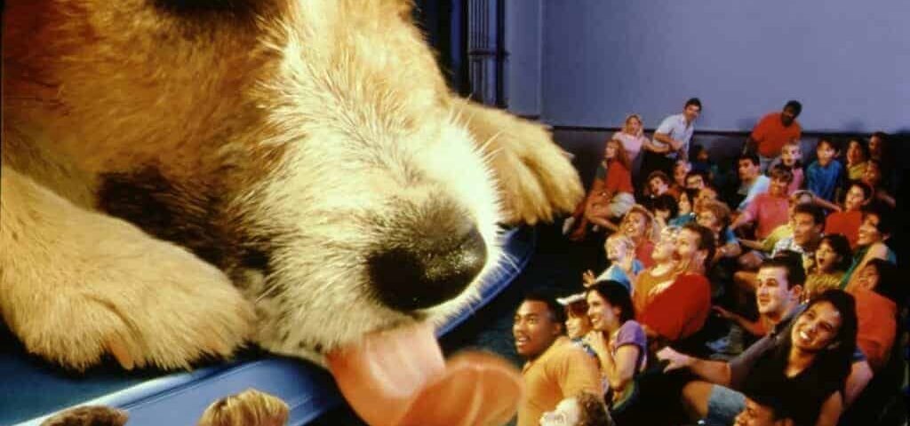Un grand gros plan d'une tête de chien superposée au premier plan, la langue sortie, surplombant un public de cinéma composé de personnes diverses riant et appréciant "Chérie, j'ai rétré
