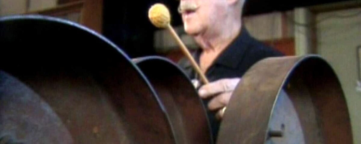 Jim MacDonald, un homme âgé portant des lunettes, joue d'un grand instrument à tambour en acier avec un maillet, visiblement engagé et appréciant sa performance.