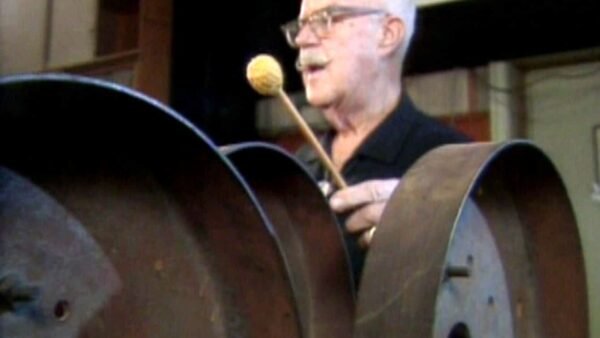 Jim MacDonald, un homme âgé portant des lunettes, joue d'un grand instrument à tambour en acier avec un maillet, visiblement engagé et appréciant sa performance.
