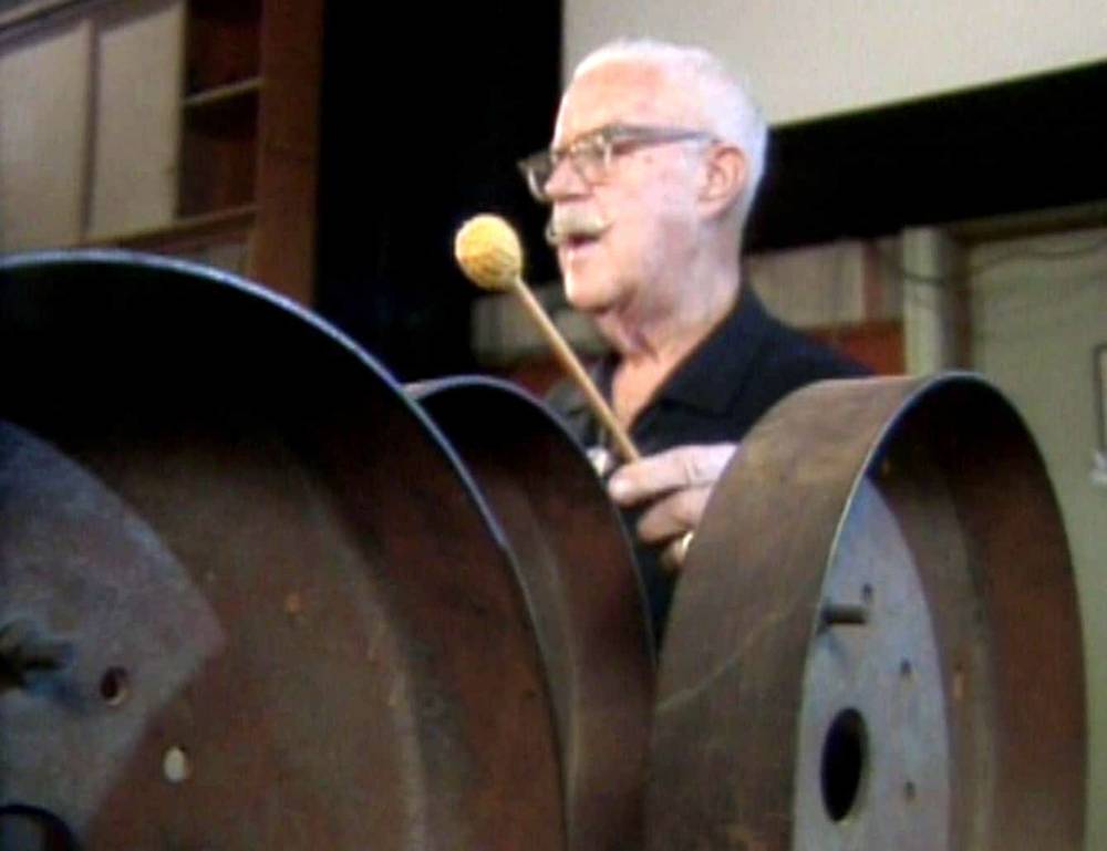 Jim MacDonald, un homme âgé portant des lunettes, joue d'un grand instrument à tambour en acier avec un maillet, visiblement engagé et appréciant sa performance.