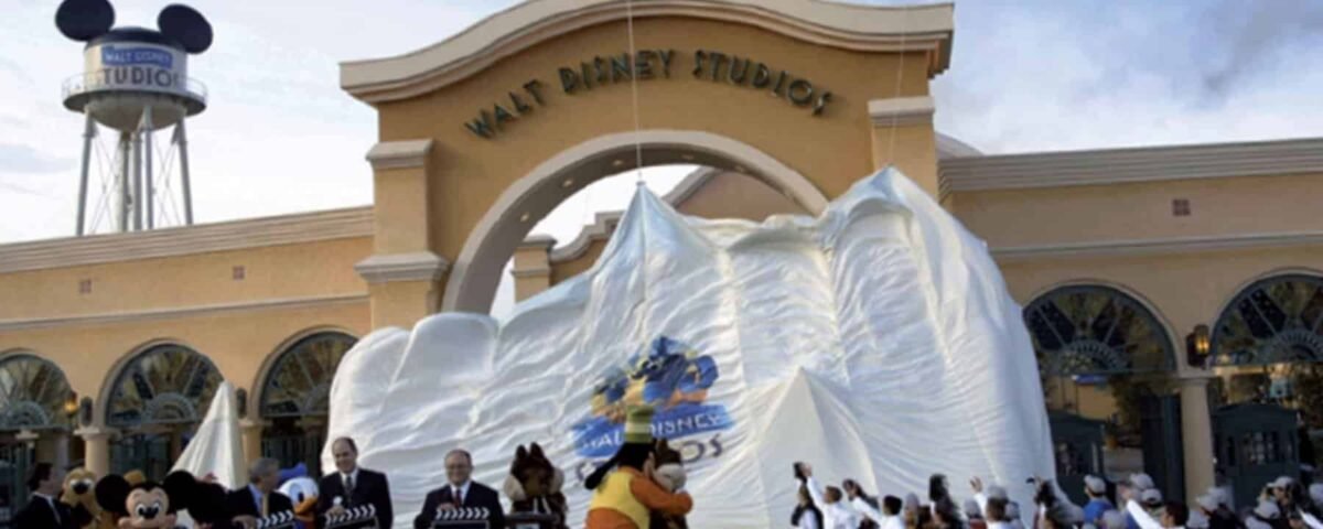 Un groupe de personnes, dont des individus en tenue de chef, se rassemblent autour d'un gros gâteau en forme de montagne à l'entrée des studios Walt Disney. Mickey Mouse est présent et fait la fête avec le groupe.