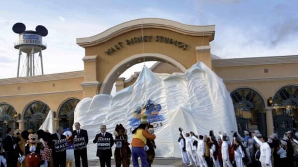 Un groupe de personnes, dont des individus en tenue de chef, se rassemblent autour d'un gros gâteau en forme de montagne à l'entrée des studios Walt Disney. Mickey Mouse est présent et fait la fête avec le groupe.