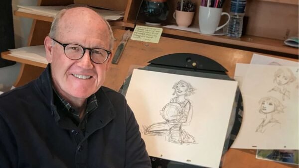 Un homme âgé portant des lunettes et une chemise noire sourit à la caméra, assis à un bureau avec des croquis de Glen Keane représentant une jeune femme exposés devant lui.