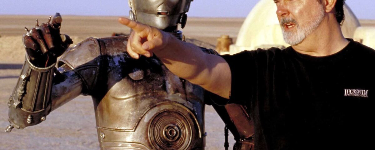 George Lucas, vêtu d'un t-shirt noir, met en scène une scène avec un personnage de robot métallique dans un vaste paysage désertique. Le robot semble conversationnel, la main tendue comme pour faire valoir un point.