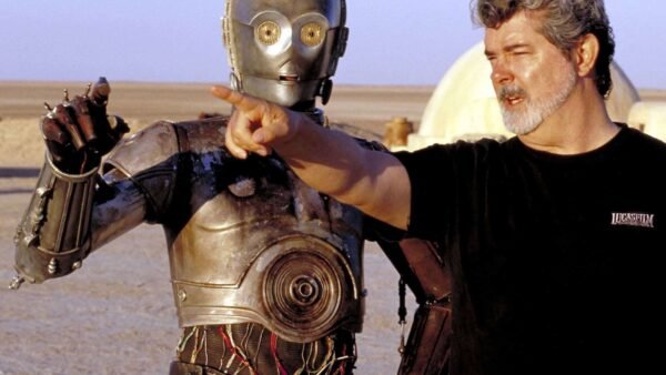 George Lucas, vêtu d'un t-shirt noir, met en scène une scène avec un personnage de robot métallique dans un vaste paysage désertique. Le robot semble conversationnel, la main tendue comme pour faire valoir un point.
