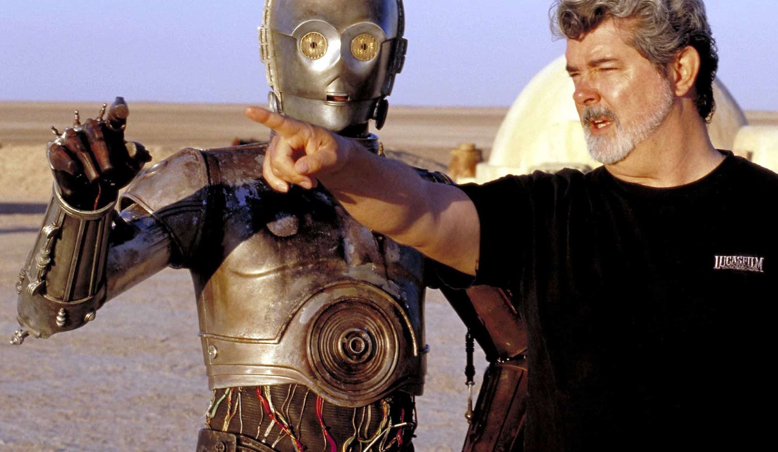 George Lucas, vêtu d'un t-shirt noir, met en scène une scène avec un personnage de robot métallique dans un vaste paysage désertique. Le robot semble conversationnel, la main tendue comme pour faire valoir un point.