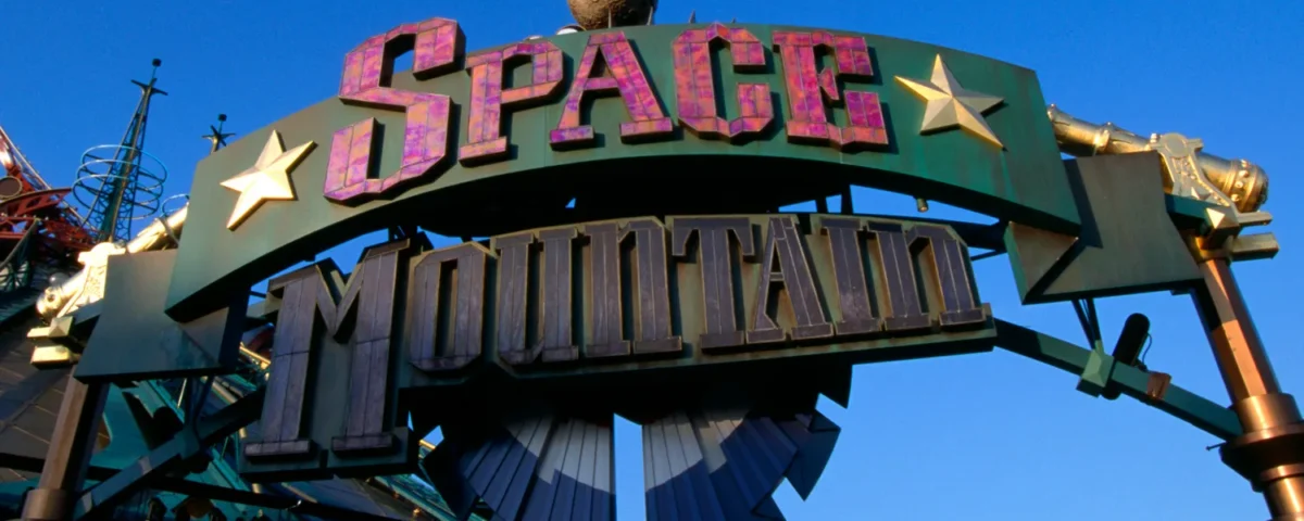 Panneau d'entrée du Space Mountain de la Terre à la Lune, comportant des lettres audacieuses et étoilées sur un ciel bleu clair, avec une partie de la structure futuriste du manège visible en arrière-plan.
