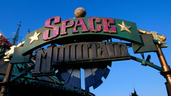 Panneau d'entrée du Space Mountain de la Terre à la Lune, comportant des lettres audacieuses et étoilées sur un ciel bleu clair, avec une partie de la structure futuriste du manège visible en arrière-plan.