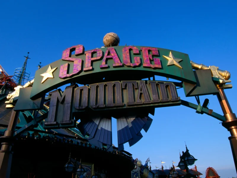 Panneau d'entrée du Space Mountain de la Terre à la Lune, comportant des lettres audacieuses et étoilées sur un ciel bleu clair, avec une partie de la structure futuriste du manège visible en arrière-plan.