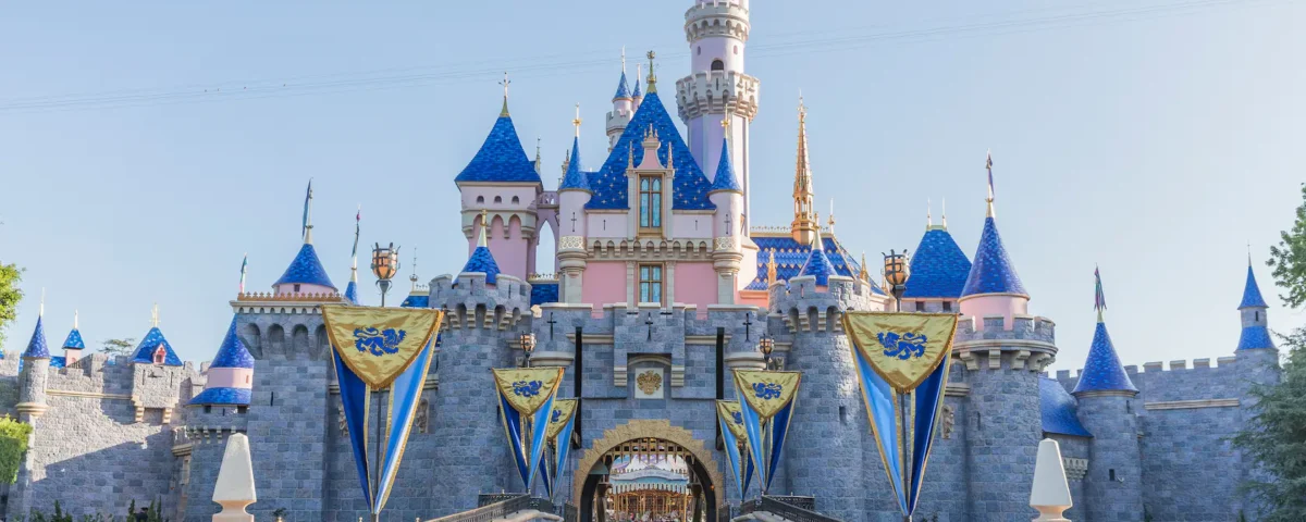 Image du château de la Belle au bois dormant à Disneyland, avec ses tourelles roses et bleues emblématiques, ornées de drapeaux dorés sous un ciel bleu clair.