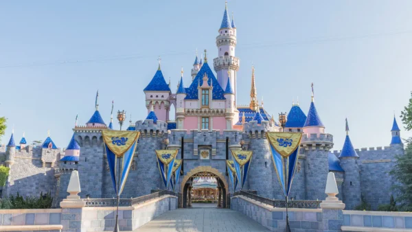 Image du château de la Belle au bois dormant à Disneyland, avec ses tourelles roses et bleues emblématiques, ornées de drapeaux dorés sous un ciel bleu clair.