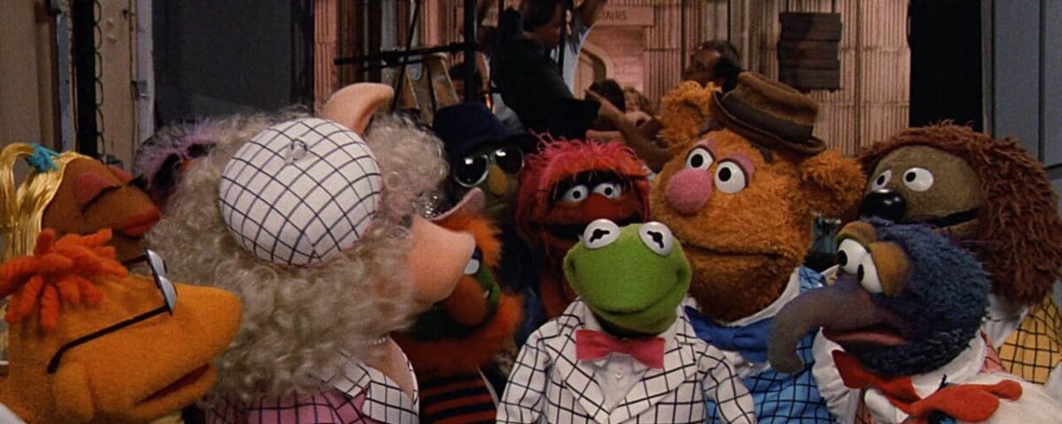 Un groupe de personnages des Muppets, dont Kermit et Miss Piggy, vêtus de diverses tenues, sont engagés dans une discussion animée dans une salle colorée et animée.
