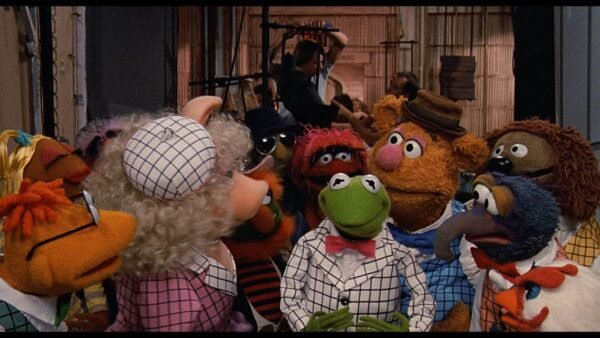 Un groupe de personnages des Muppets, dont Kermit et Miss Piggy, vêtus de diverses tenues, sont engagés dans une discussion animée dans une salle colorée et animée.