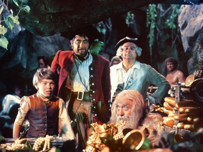 Un groupe d'aventuriers, dont un jeune garçon, un homme robuste et un homme plus âgé, examinent un trésor sur "l'Île au Trésor" dans une grotte remplie de pièces d'or.