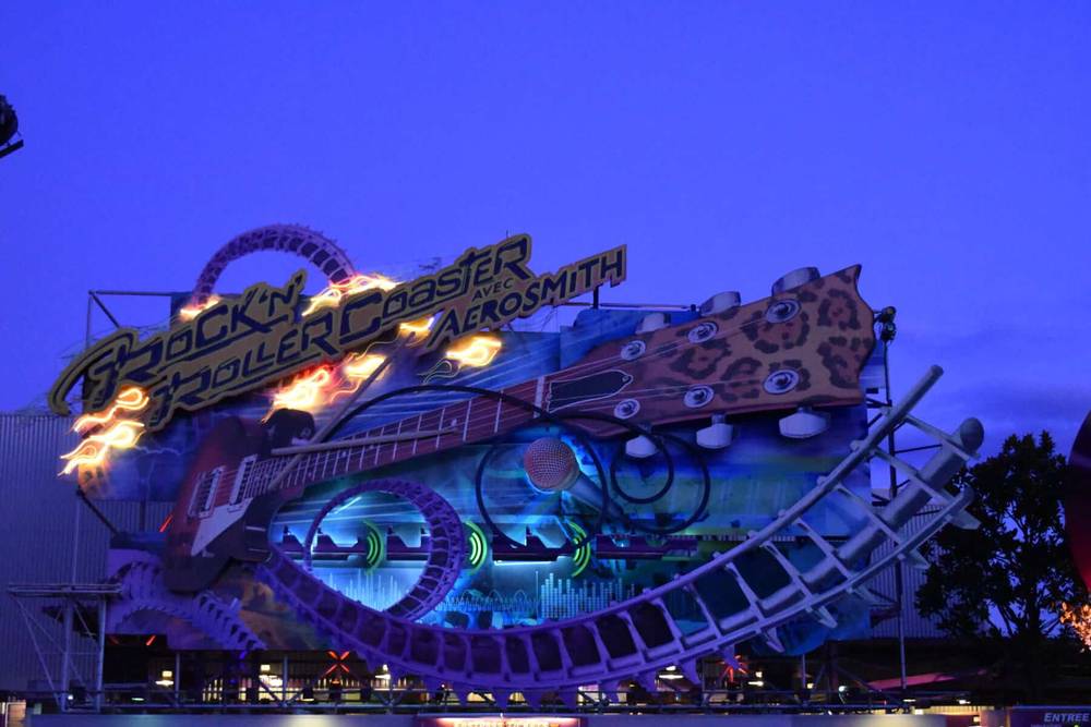 Entrée éclairée au néon du Rock 'n' Roller Coaster mettant en vedette Aerosmith au crépuscule, affichant une grande piste de montagnes russes en boucle et des éléments musicaux.