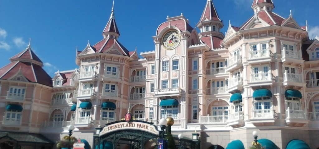 L'entrée de Disneyland présente un palais orné de façades roses et blanches, de toits pointus et d'une horloge centrale, sous un ciel bleu clair.