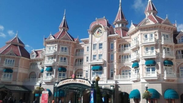 L'entrée de Disneyland présente un palais orné de façades roses et blanches, de toits pointus et d'une horloge centrale, sous un ciel bleu clair.