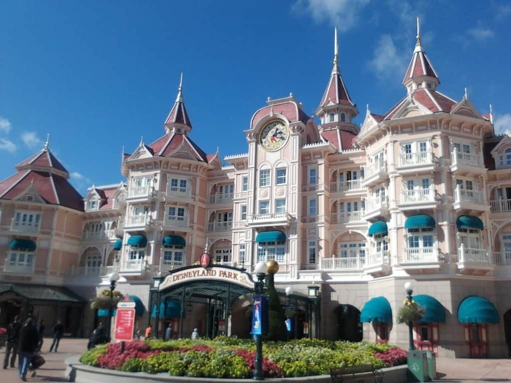 L'entrée de Disneyland présente un palais orné de façades roses et blanches, de toits pointus et d'une horloge centrale, sous un ciel bleu clair.