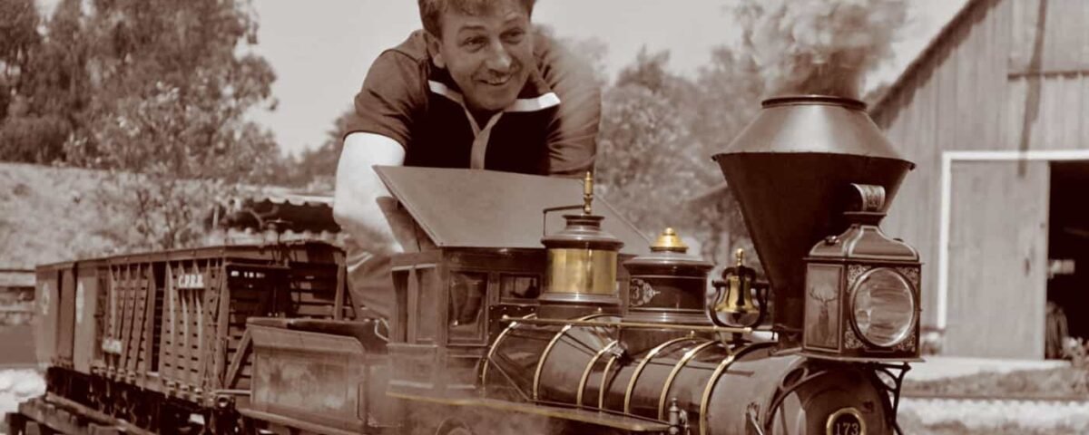 Un homme souriant tout en se penchant depuis la cabine d'une locomotive à vapeur d'époque, qui circule sur des rails à voie étroite dans une scène sépia. Une grange est visible en arrière-plan.