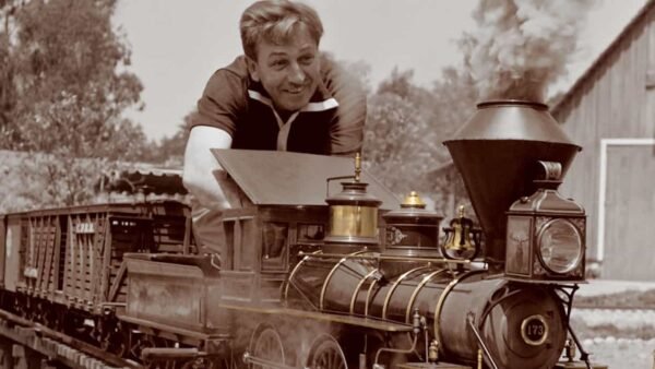 Un homme souriant tout en se penchant depuis la cabine d'une locomotive à vapeur d'époque, qui circule sur des rails à voie étroite dans une scène sépia. Une grange est visible en arrière-plan.