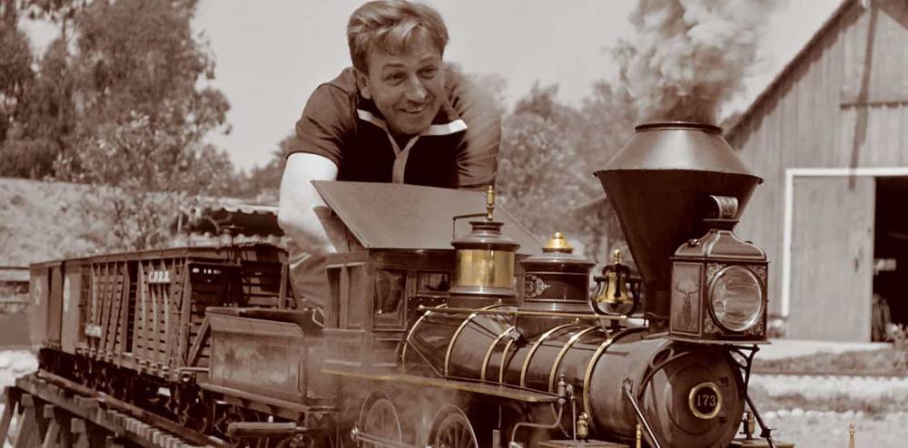 Un homme souriant tout en se penchant depuis la cabine d'une locomotive à vapeur d'époque, qui circule sur des rails à voie étroite dans une scène sépia. Une grange est visible en arrière-plan.