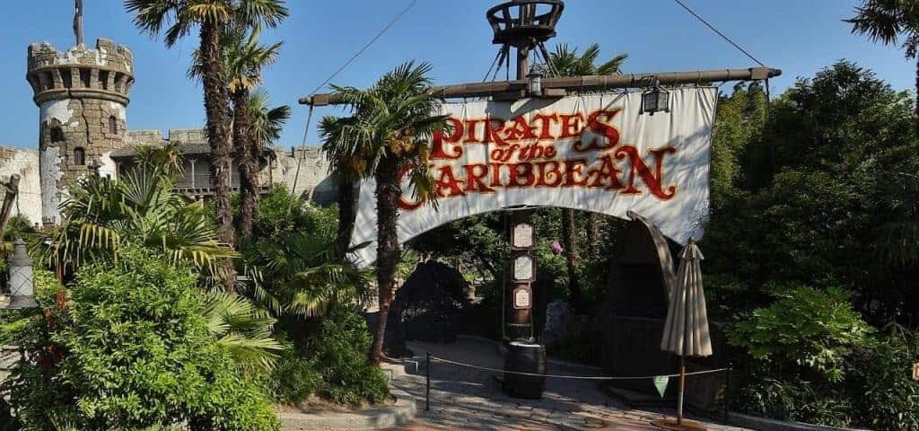 Entrée du manège "Pirates des Caraïbes" à Adventureland, avec un panneau sur le thème des pirates, des plantes vertes luxuriantes et une tourelle de château en arrière-plan.