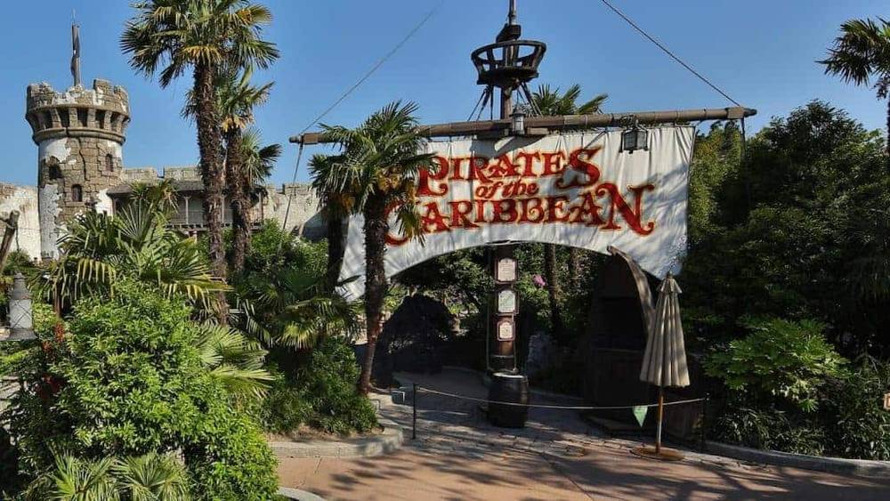 Entrée du manège "Pirates des Caraïbes" à Adventureland, avec un panneau sur le thème des pirates, des plantes vertes luxuriantes et une tourelle de château en arrière-plan.