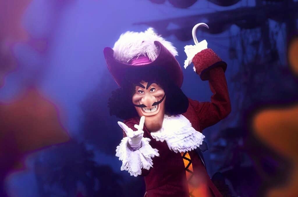 Une personne habillée en Capitaine Crochet, posant avec un geste de la main et un sourire narquois, portant un grand chapeau rouge, un manteau rouge foncé à volants blancs et tenant un crochet dans l'autre
