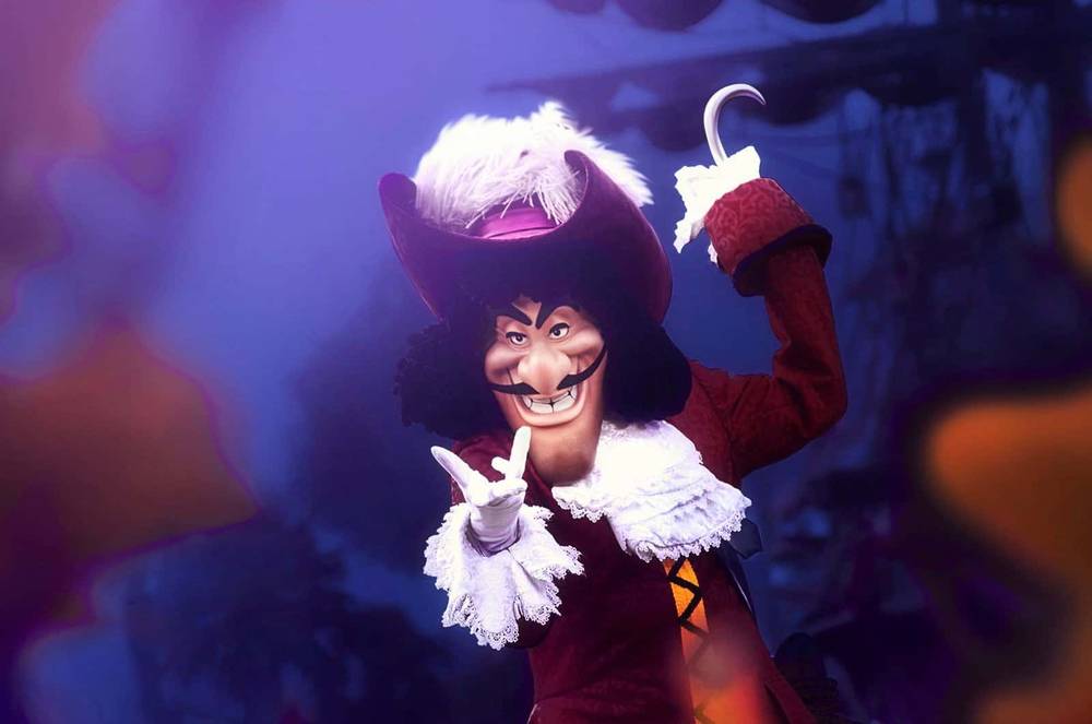 Une personne habillée en Capitaine Crochet, posant avec un geste de la main et un sourire narquois, portant un grand chapeau rouge, un manteau rouge foncé à volants blancs et tenant un crochet dans l'autre