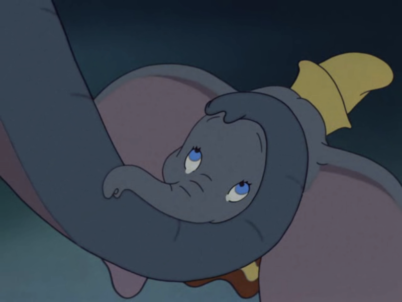 Une image de dessin animé de Dumbo, le petit éléphant des chansons de Disney, l'air triste et reposant sa tête et sa trompe sur ses jambes croisées, avec un petit chapeau jaune sur la tête.
