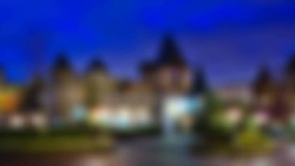 L'image est une vue floue d'une rue bordée de maisons pittoresques sous un ciel crépusculaire à Disneyland Paris, les lumières des bâtiments projetant une douce lueur sur les environs.