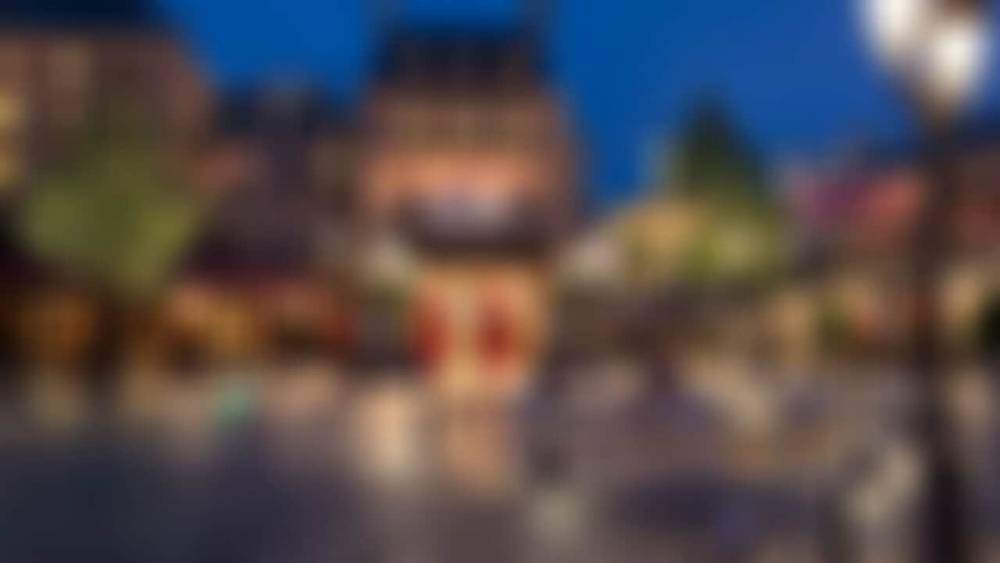 Vue floue d'une place animée de Disneyland Paris au crépuscule, avec des bâtiments colorés et des lampadaires lumineux projetant une lueur chaleureuse.