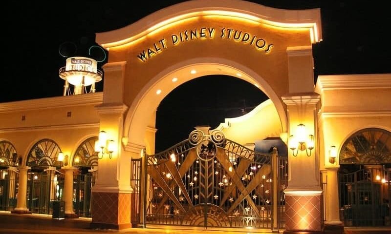 Vue nocturne de la porte d'entrée éclairée de Disneyland Paris, avec une grande arche portant le nom du studio et une porte ornée en dessous.