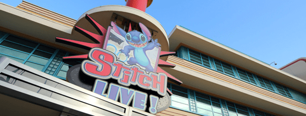 Coloré « Stitch Live ! » panneau représentant le personnage animé Stitch, monté sur la façade d'un immeuble moderne sous un ciel bleu.