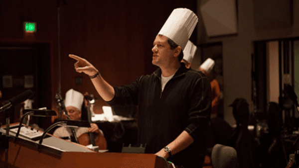 Un chef au chapeau blanc se tient sur un podium, pointant du doigt, avec d'autres chefs assis derrière lui dans une pièce faiblement éclairée, discutant des nuances de la ratatouille.