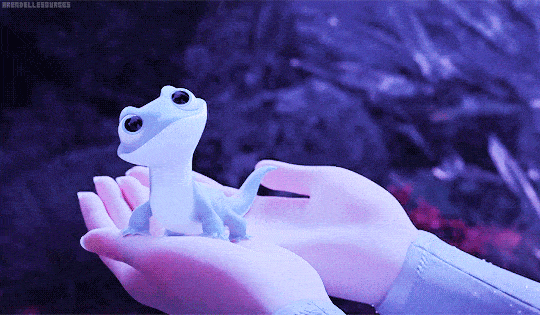 Une image animée montrant un petit gecko blanc aux yeux écarquillés assis sur une main humaine, avec un arrière-plan violet légèrement flou de plantes du film Frozen.