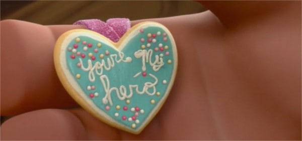 Gros plan d'une main tenant un biscuit en forme de cœur décoré d'un glaçage bleu sarcelle, de pépites roses et blanches, et des mots « tu es mon héros » écrits en cursive avec un glaçage rose inspiré de