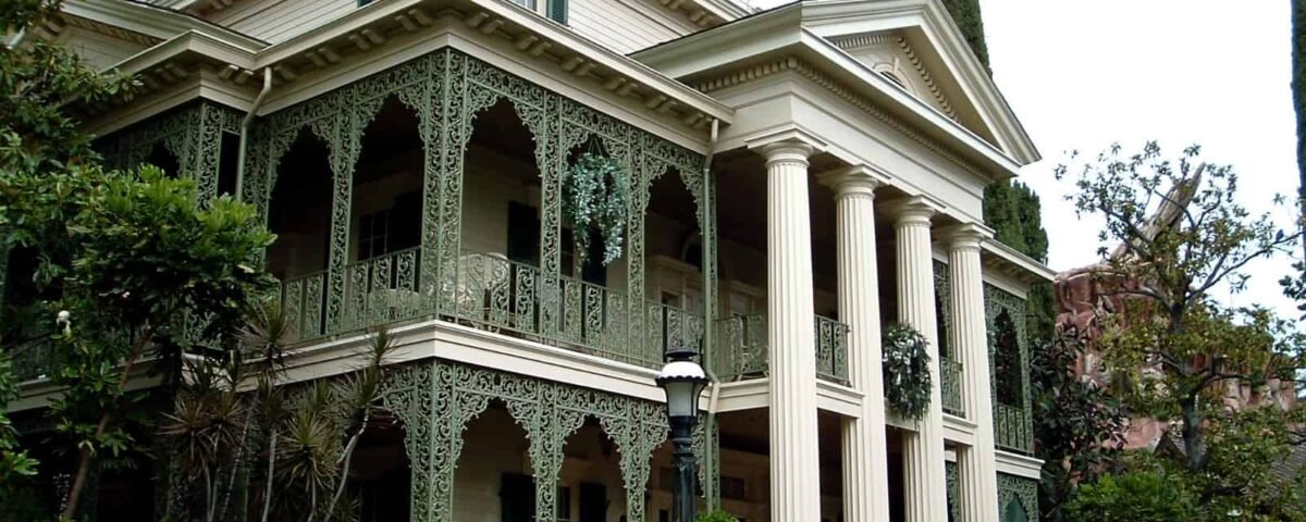 Un grand manoir richement conçu avec de hautes colonnes blanches, des ferronneries vertes complexes sur les balcons et entouré d'une verdure luxuriante et d'arbres, rappelant l'attraction de maison hantée Phantom Manor à Disneyland Paris.