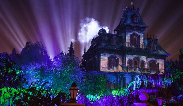 Un Phantom Manor illuminé la nuit avec des faisceaux de lumière se projetant dans le ciel, entouré d'arbres étrangement éclairés et d'une ambiance brumeuse.