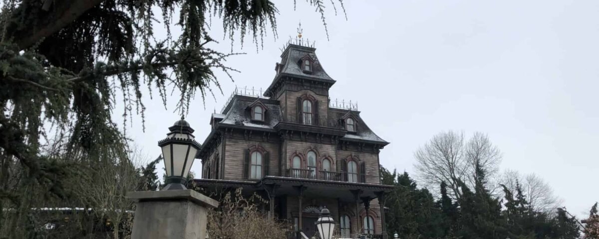 Un manoir fantôme de style victorien dans un décor hivernal, entouré d'arbres dénudés et d'une légère couche de neige, sous un ciel couvert.