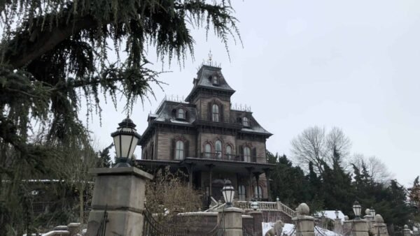 Un manoir fantôme de style victorien dans un décor hivernal, entouré d'arbres dénudés et d'une légère couche de neige, sous un ciel couvert.