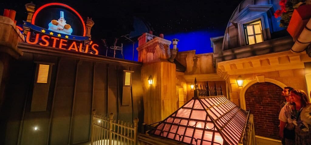 Une scène nocturne animée à Disneyland Paris avec l'enseigne lumineuse « Gusteau's » au-dessus d'une façade de style français, avec deux visiteurs levant les yeux avec admiration.